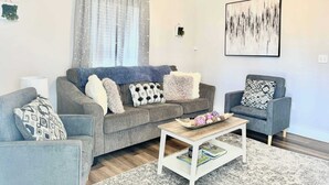 A living room with a grey sofa, a coffee table, and a grey armchair. There is a painting on the wall and a window with curtains. - Newly Remodeled 2 Bedroom Home in Shelbyville! (Shelbyville)