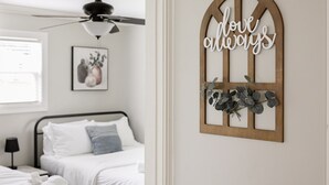 Un dormitorio con cama, ventilador de techo, un cuadro enmarcado y un letrero decorativo en la pared.