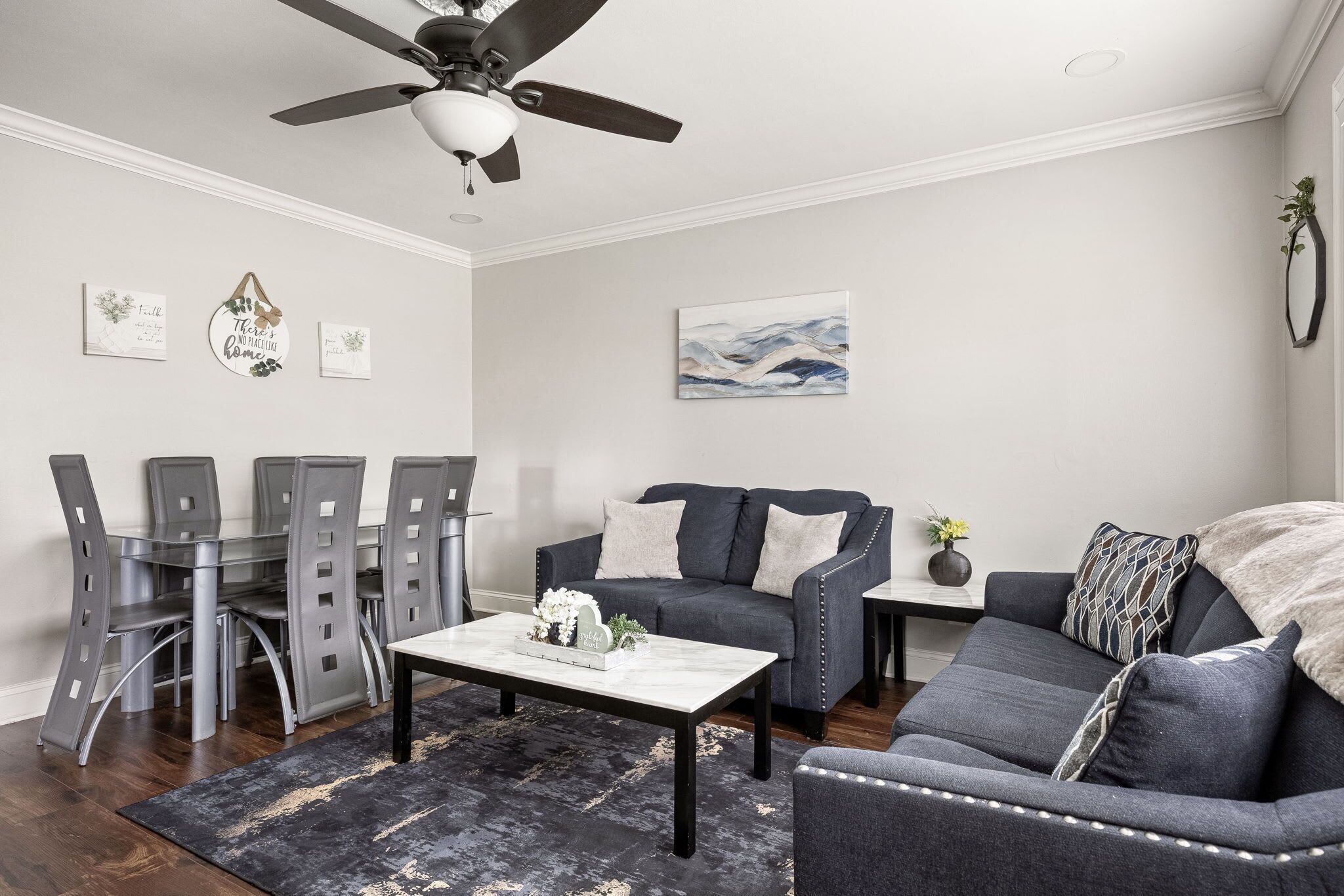 A modern living room with a dark gray sofa, a glass dining table, and a ceiling fan.