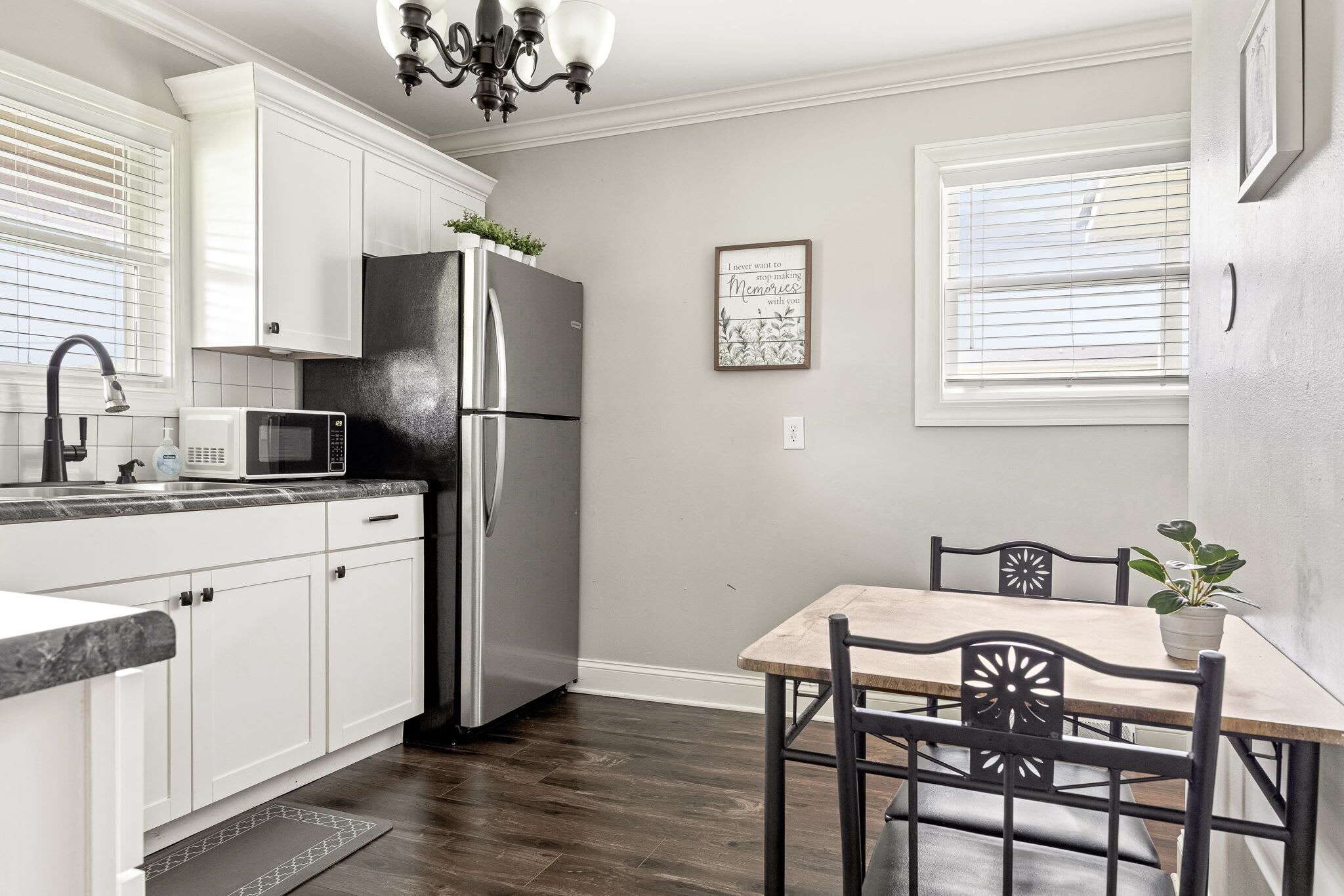 A kitchen with white cabinets, a stainless steel refrigerator, a microwave, a dining table with chairs, and a potted plant.