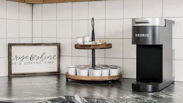 Una cocina con cafetera, una bandeja con tazas de café y un letrero enmarcado.