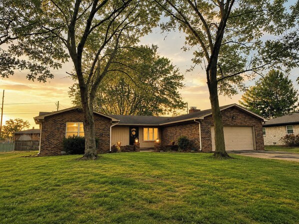 Une maison à deux étages, construite en briques, avec un garage à l’avant, entourée d’une pelouse bien entretenue et de grands arbres.