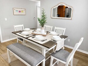 Een eethoek met een houten tafel voor zes personen, witte stoelen, een bank en een spiegel aan de muur.