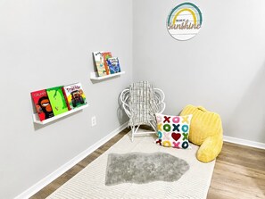 Een kamer met een witte wand, een witte stoel, een gele relaxfauteuil, een grijze vloerkleed en kinderboeken op een witte plank.