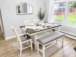 A dining area with a table set for four, white chairs, a bench, a mirror, and a potted plant.