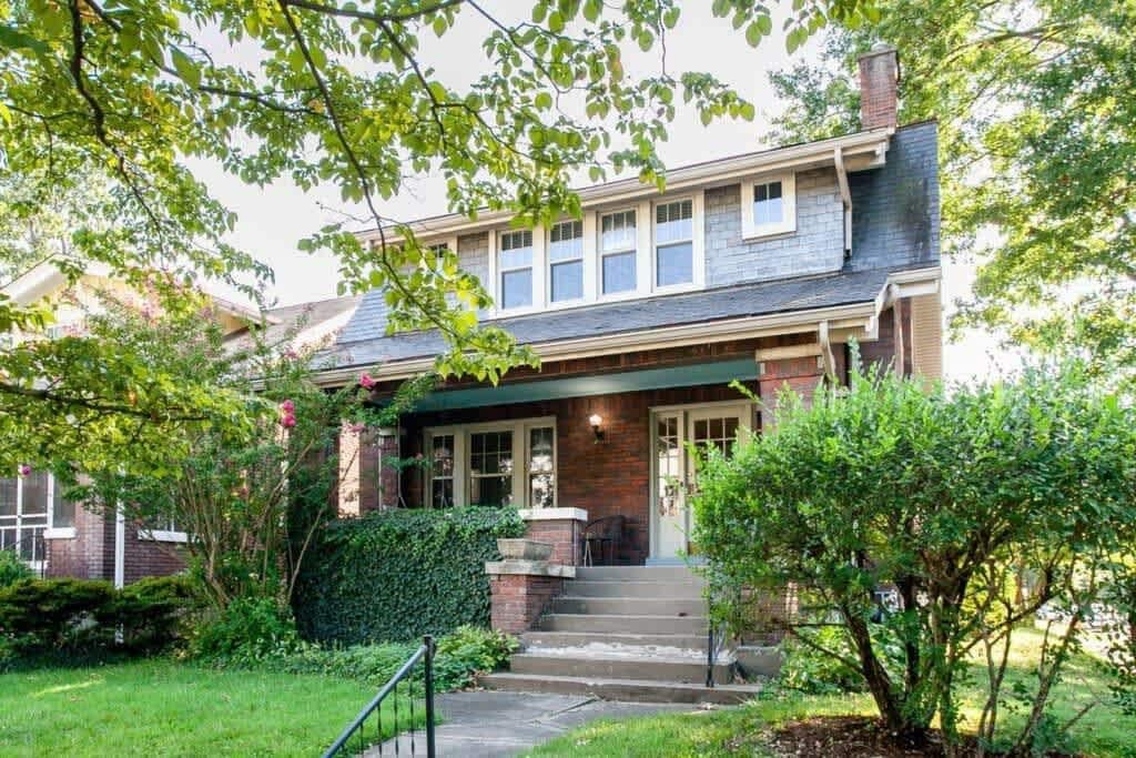 Une maison à deux étages avec un porche à l’avant, une façade en briques et un petit jardin devant, agrémenté d’un arbre et de quelques arbustes.