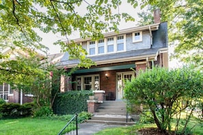 A two-story house with a front porch, brick exterior, and a small front yard with a tree and shrubbery. - Highlands Haven | Walkable | Charming Area (Louisville)