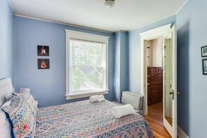 A bedroom with a large bed, a window with blinds, a door leading to a hallway, and a small bedside table. - Highlands Haven | Walkable | Charming Area (Louisville)