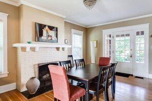 A dining room with a brick fireplace, a dining table with chairs, and a large window with blinds.
