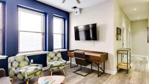 A modern living room with a blue accent wall, a flat-screen TV mounted on the wall, a wooden desk, two patterned armchairs, and a small coffee table. - Theatre District Condo | Gym • Free Parking • DT (Louisville)