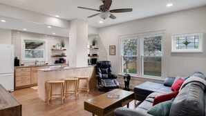 Una cocina moderna con una isla central, una zona de comedor con una mesa de madera y taburetes altos, un sofá de cuero con cojines y un ventilador de techo.