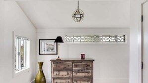 Una habitación con piso de madera, un aparador de estilo antiguo, un jarrón verde, un cuadro enmarcado, una lámpara negra y un ventanal decorativo con vidrieras.