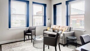A modern living room with a grey sofa, two armchairs, a coffee table, and a side table. There are three windows with blinds, and a floor lamp is visible. - Modern 2BR Luxury Condo • Downtown w/ Garage & Gym (Louisville)