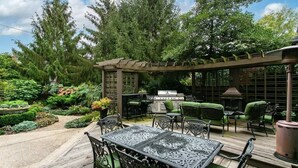 Un patio con una pérgola, mobiliario exterior y zona de barbacoa, rodeado de abundante vegetación y árboles.