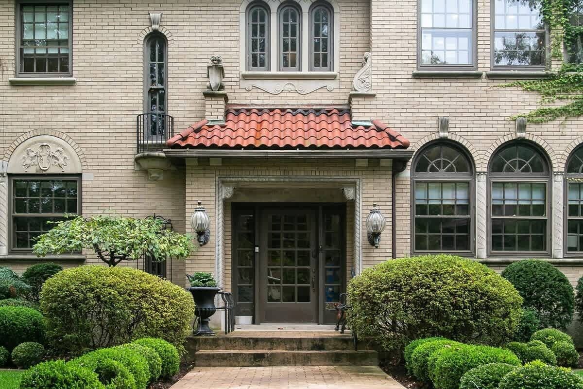 A stately building with a symmetrical facade, a central entrance with a red-tiled roof, and a well-manicured garden with hedges and a tree.