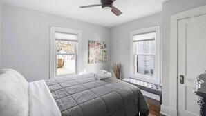 A bedroom with a bed, a ceiling fan, two windows, a bench, and a door.