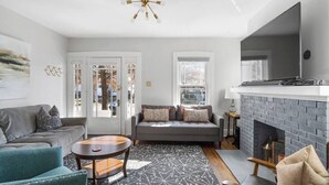 A modern living room with a gray sofa, a teal armchair, and a round coffee table. There is a fireplace with a brick wall and a flat-screen TV mounted above it.