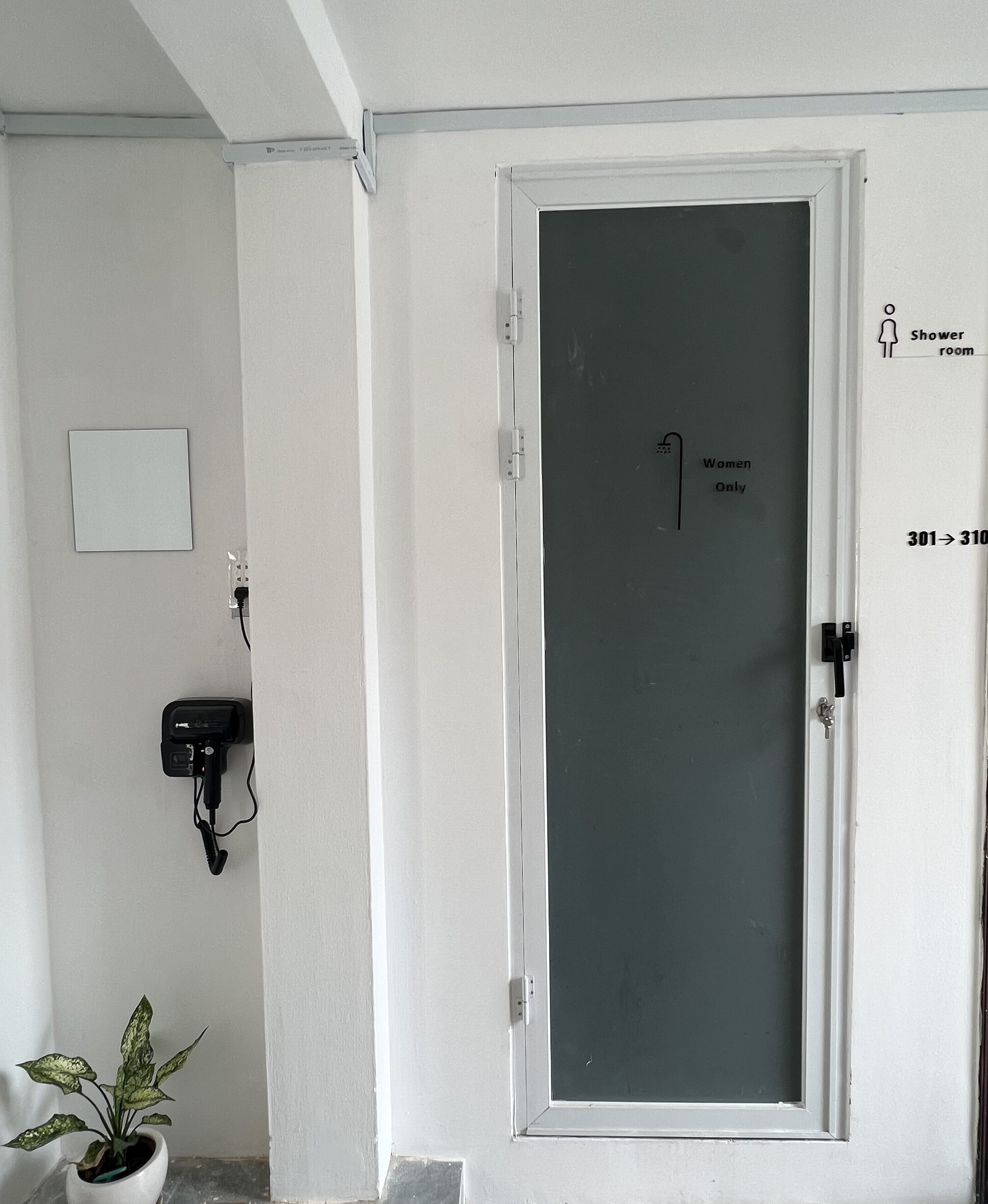A white door with a frosted glass panel, a small potted plant, a power outlet, and a sign indicating a women's restroom and shower room.