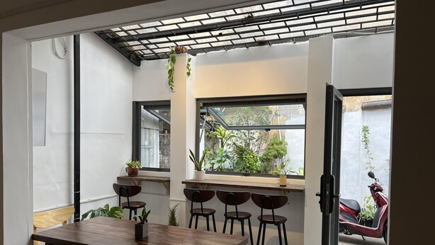 A dining area with a wooden table and chairs, a window with a view of the outside, and a ceiling with a grid pattern. - 8h - Hostel  (Ho Chi Minh City)