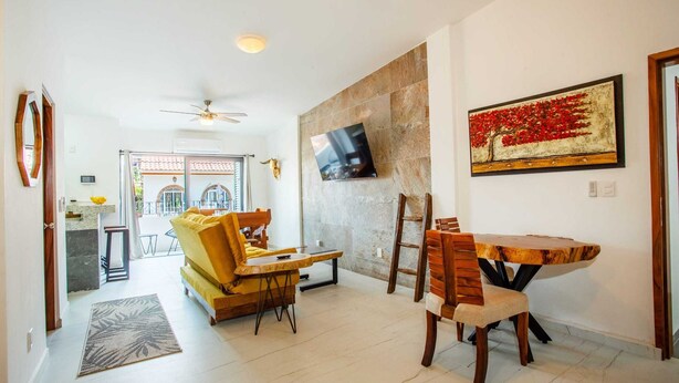 A spacious living area with a sofa, dining table, and chairs, a flat-screen TV mounted on a stone wall, and a view into the kitchen. - Beautiful Casa Madero 470 (Puerto Vallarta)