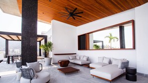 Una sala de estar moderna con un sofá seccional grande, una mesa de centro y un espejo amplio. El techo es de madera y hay un ventanal con vistas a palmeras.