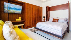 Un dormitorio con cabecera de madera, cama con ropa de cama a rayas, sofá amarillo, televisor montado en un panel de madera y un cuadro de una playa.