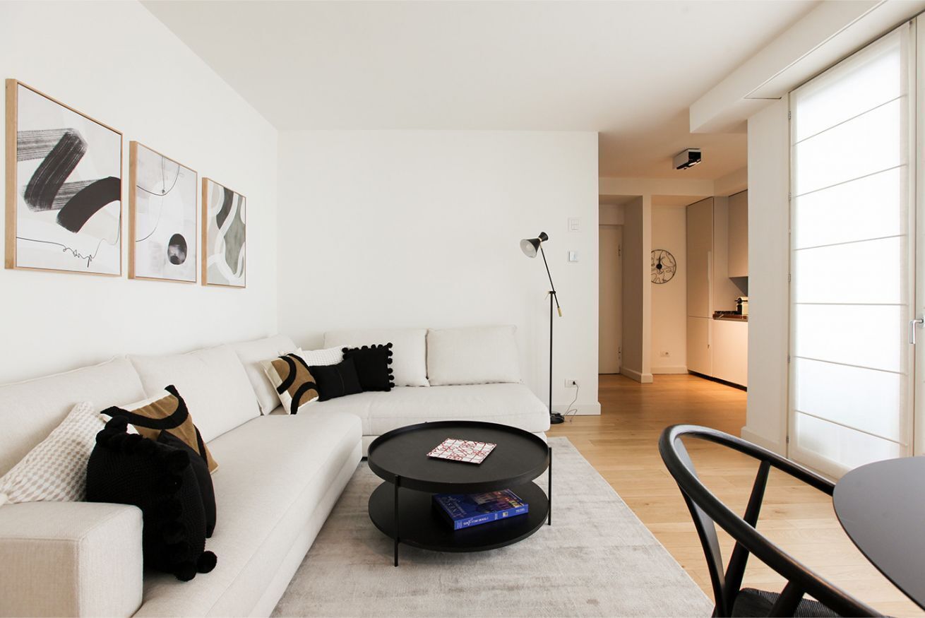 Un salon moderne avec un canapé blanc, une table basse noire et un coin repas avec une chaise. Des tableaux abstraits ornent le mur, et une horloge y est également accrochée.