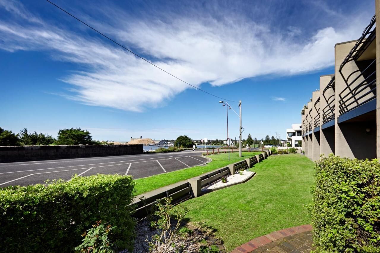 A parking lot with a clear sky, a grassy area, and a building with balconies.