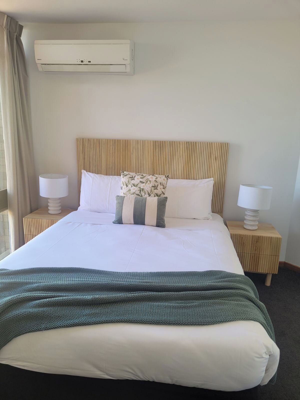 A hotel room with a large bed, two bedside tables, a wall-mounted air conditioner, and a window with curtains.