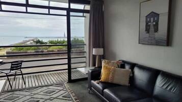Een moderne woonkamer met een leren zwarte bank, een getextureerd vloerkleed en uitzicht op zee door grote ramen.