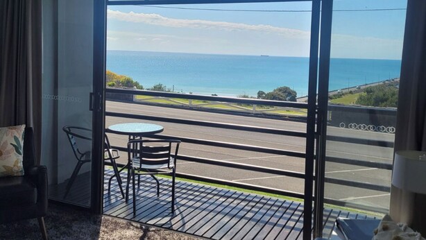 A room with a large sliding glass door leading to a balcony with a view of the sea and a road. - Centreport Units (Portland)