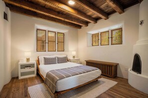 Un dormitorio con cama de madera, paredes blancas y techo de madera. Hay dos ventanas con contraventanas de madera, una mesita de noche con lámpara y un estante pequeño.