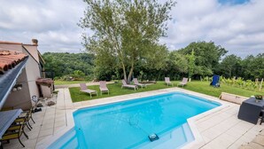 A rectangular swimming pool with a tiled deck, surrounded by a grassy area with lounge chairs and a tree.