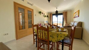 A dining area with a table set for four, surrounded by wooden chairs. There is a sofa in the background and a window with curtains.