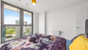 Camera d'albergo moderna con ampie finestre, un letto con lenzuola a motivi floreali, una coperta arrotolata e un cuscino giallo.