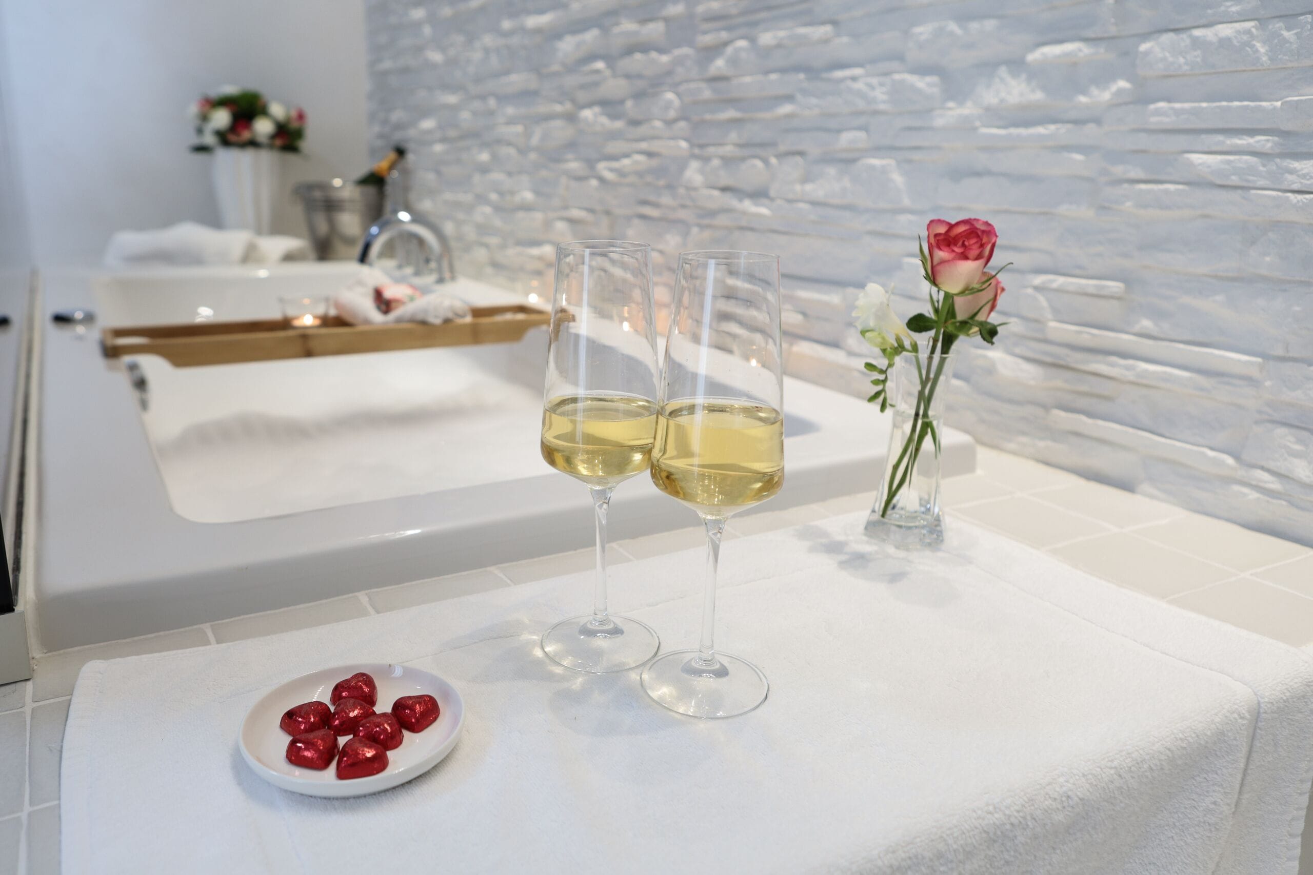 A clean, modern bathroom with a white marble countertop, two glasses of champagne, a small plate of chocolates, and a vase with a single rose.