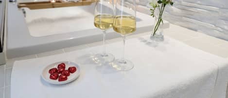 A clean, modern bathroom with a white marble countertop, two glasses of champagne, a small plate of chocolates, and a vase with a single rose.