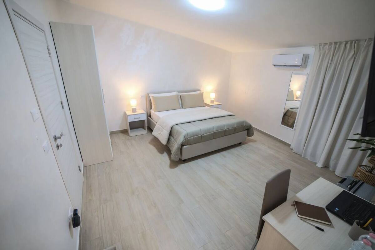 A bedroom with a bed, bedside tables, a desk with a laptop, and an air conditioning unit.