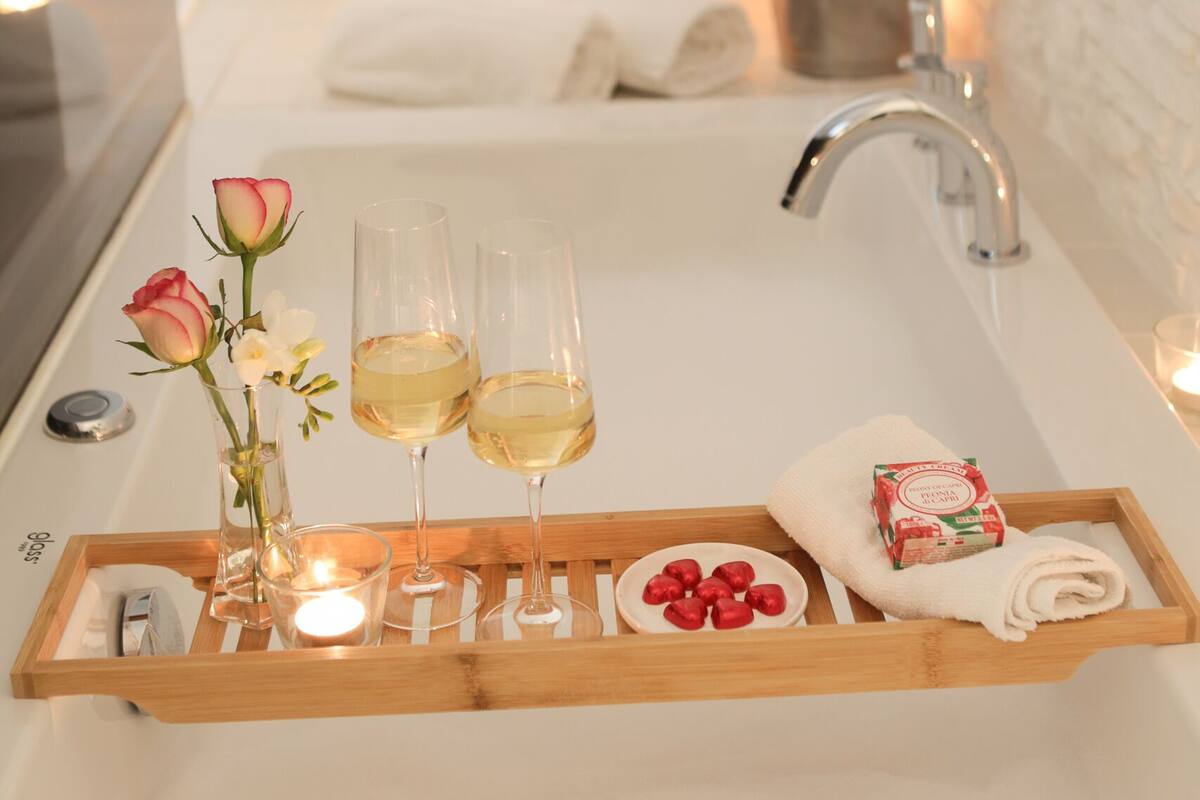 A bathroom with a wooden tray holding two glasses of champagne, a vase with flowers, and a bowl of strawberries.