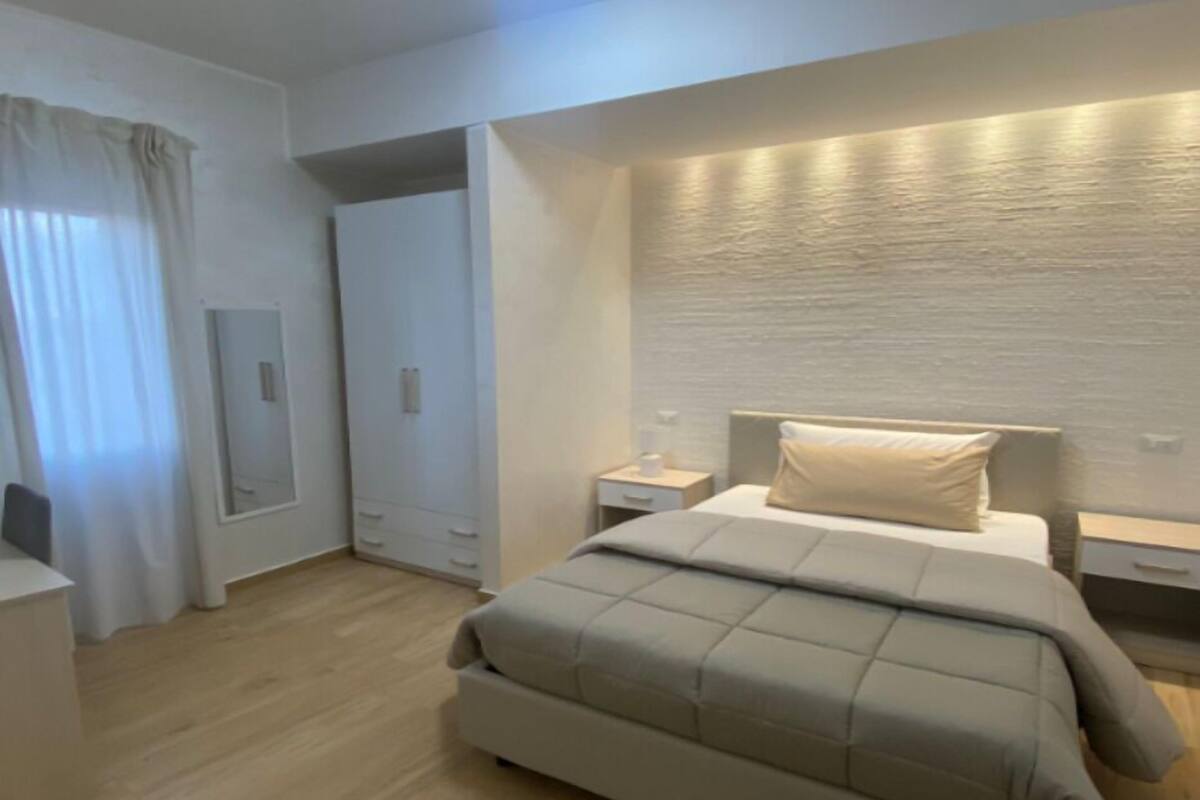 A modern bedroom with a large bed, a nightstand, a bedside table, a wardrobe, and a mirror.