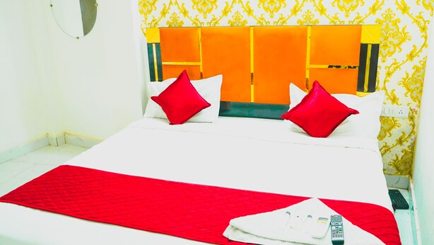 A hotel room with a large bed, two red pillows, a red blanket, a white pillow, and a folded white towel with a remote control on it. - Roomshala 080 Hotel Kings (New Delhi)