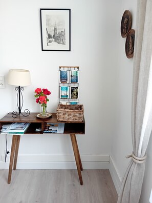 A wooden desk with a lamp, a vase with flowers, a wicker basket, and a framed picture of a town.