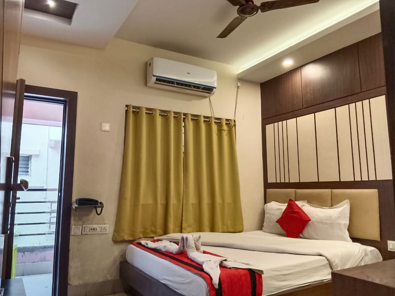 A hotel room with a bed, a ceiling fan, an air conditioning unit, a telephone, and a window with curtains.