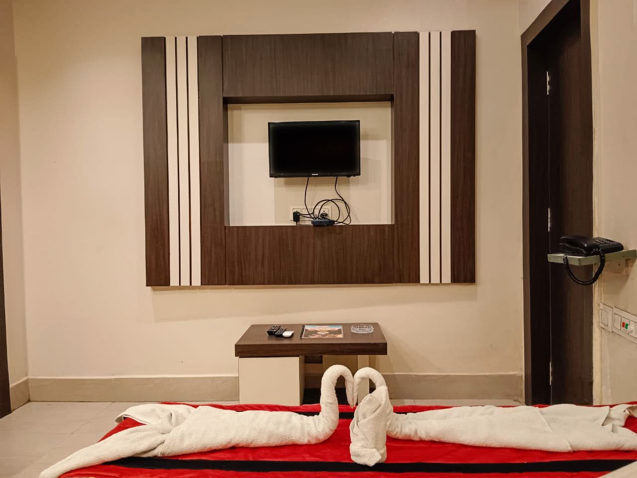 A hotel room with a flat-screen TV mounted on the wall, a small table with a remote control and a magazine, and a red and black striped bedspread.