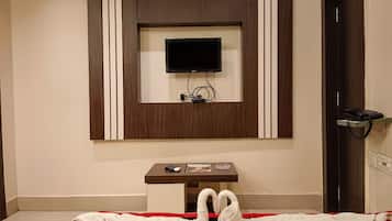A hotel room with a flat-screen TV mounted on the wall, a small table with a remote control and a magazine, and a red and black striped bedspread.