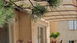 An outdoor patio with a dining table and chairs, surrounded by a pergola and a small tree.