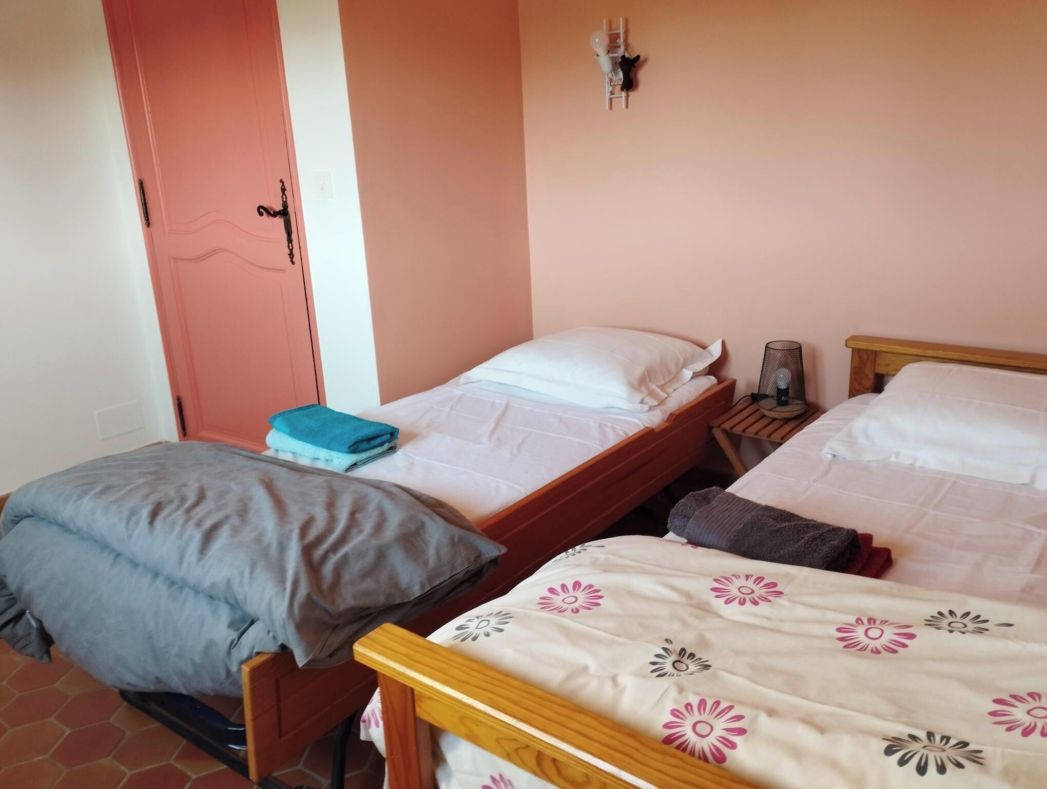 Una habitación con dos camas, una con colcha estampada y la otra lisa. Hay una mesita con una lámpara y una silla, y una puerta rosa.