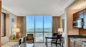 A hotel room with a large window, a dining area, a sofa, a table, and a kitchenette.
