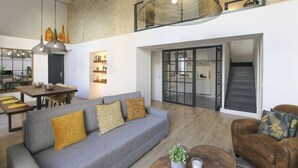 Uma sala espaçosa com um sofá cinza, uma mesa de centro de madeira e uma poltrona de couro marrom. Uma escada leva a um andar superior com uma janela grande.