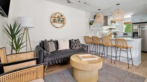 A modern living room with a grey sofa, round coffee table, and rattan chairs. A kitchen with white cabinets and a central island is visible in the background. - Beautiful Penthouse Condo Just Steps to King (Charleston)
